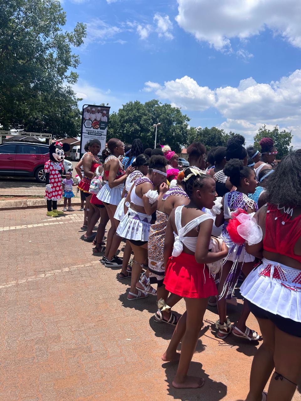 A procession of young women in traditional attire passing the Isinkwa Sethu banner